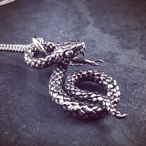 Op de afbeelding: Een zilveren slangenhangerketting met een opgerold slangendesign. De kop van de slang is omhoog en de mond is open. De ketting ligt op een donkergrijze achtergrond.