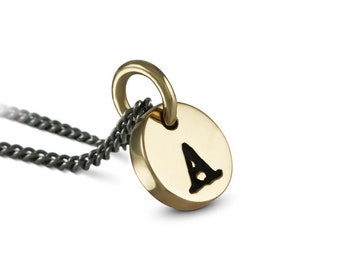 May include: A gold necklace with a small round pendant featuring the letter A in black. The chain is black and has a curb link style.
