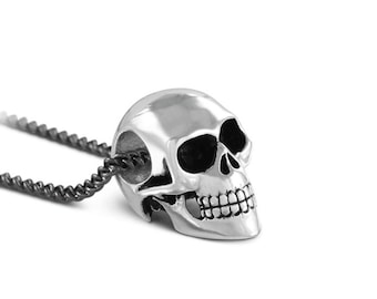 May include: A silver skull pendant necklace with a black chain. The skull is detailed with a prominent jawline and eye sockets.