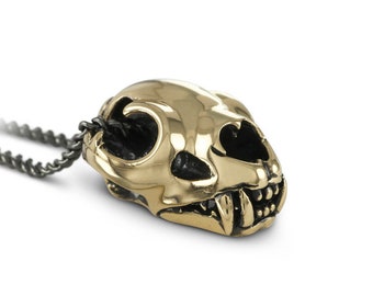 May include: A gold-colored pendant necklace featuring a detailed skull of a small cat. The pendant is attached to a black chain.