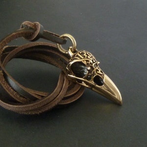 May include: A gold-tone pendant necklace featuring a detailed bird skull design with black enamel accents. The pendant is attached to a brown leather cord.