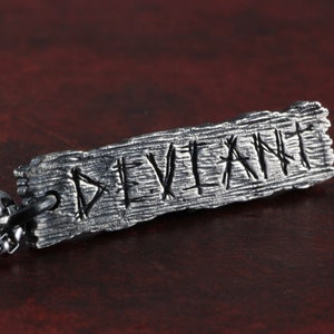 Può includere: Una collana d'argento con un ciondolo rettangolare che dice "DEVIANT" in un font stilizzato. Il ciondolo ha una superficie testurizzata che ricorda il legno.