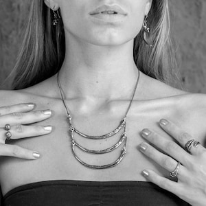 Peut inclure: Un collier en argent avec trois pendentifs en forme de croissant de lune superposés. Le collier est porté par une personne aux cheveux longs.