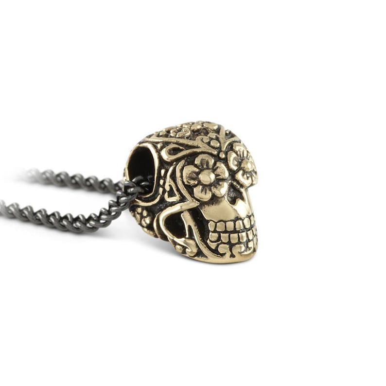May include: A gold-coloured skull pendant with floral and scrollwork details. The skull is attached to a dark grey chain. The pendant has a detailed design and is set against a white background. The jewellery is likely a necklace.
