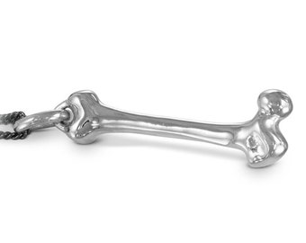 May include: A silver bone pendant on a dark chain. The pendant is a detailed, three-dimensional representation of a bone, with rounded ends and a central shaft. The chain is a dark, twisted rope style.