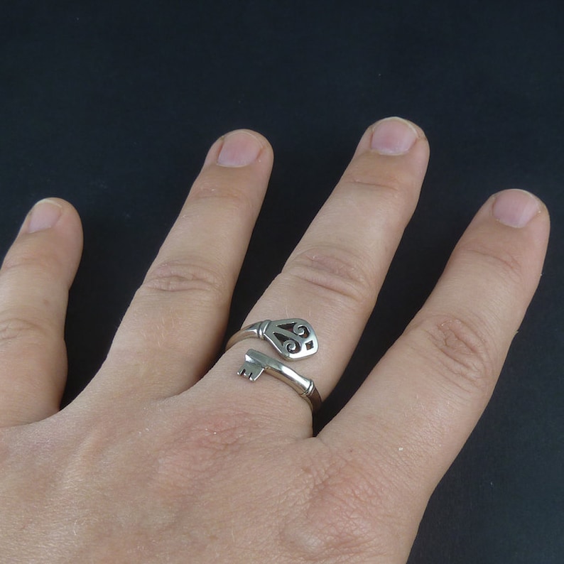 May include: A silver key-shaped ring with a decorative design on the key head. The ring is worn on a finger.