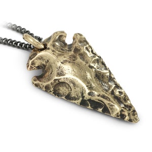 May include: A bronze arrowhead pendant on a dark chain. The arrowhead has a textured surface and a loop for the chain. The chain is dark grey. The pendant is a unique piece of jewellery.