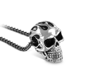 May include: A silver skull pendant necklace with a black rope chain. The skull has a flame design on the top of its head.