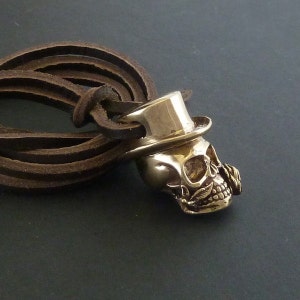 May include: A gold-toned skull pendant with a top hat and a small rose on the side. The pendant is attached to a brown leather cord.