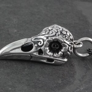 May include: A silver pendant in the shape of a crow skull with intricate floral details and black onyx eyes. The pendant has a loop for attaching to a chain.