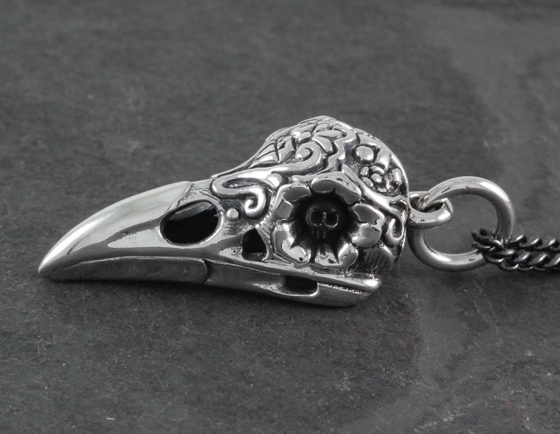 May include: A silver pendant in the shape of a crow skull with intricate floral details and black onyx eyes. The pendant has a loop for attaching to a chain.