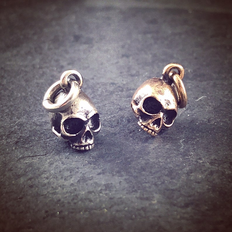 May include: Two silver skull charms with a loop for attaching to a necklace or bracelet. The skull on the left is silver, and the skull on the right is gold.