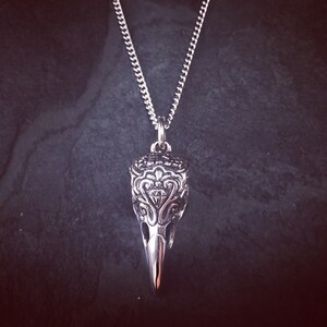 May include: A silver necklace with a detailed, ornate, bird skull pendant. The pendant has a diamond-shaped design in the center.