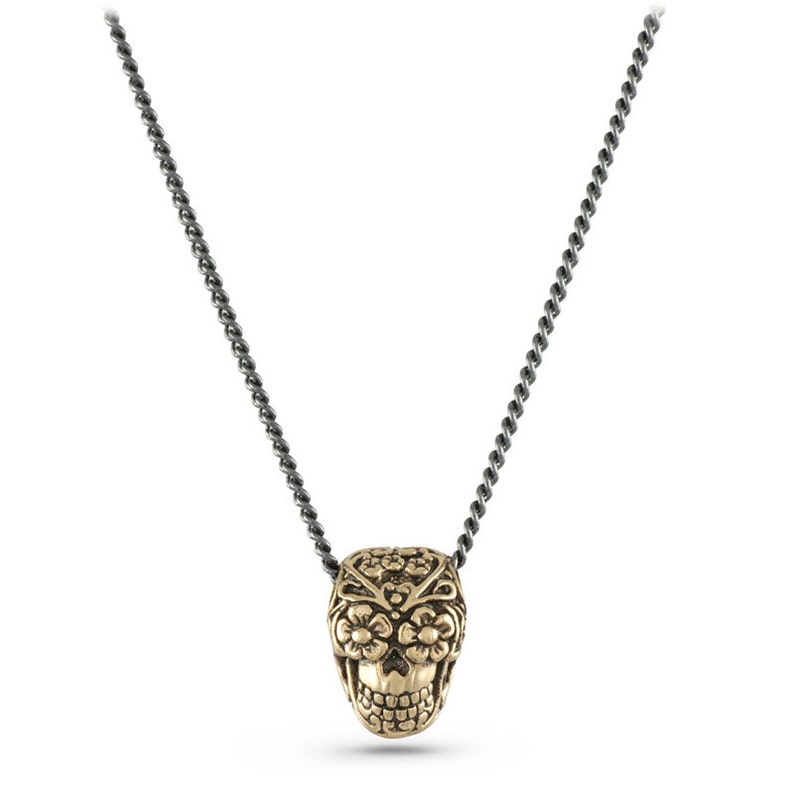 May include: A gold-coloured skull pendant necklace with a detailed floral design. The skull is suspended from a dark grey chain. The pendant features intricate carvings and a detailed skull face.