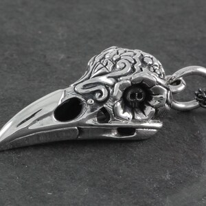 May include: Silver pendant featuring a detailed, ornate crow skull with floral designs. The skull has a small skull detail inside the eye socket. The pendant is attached to a black chain.