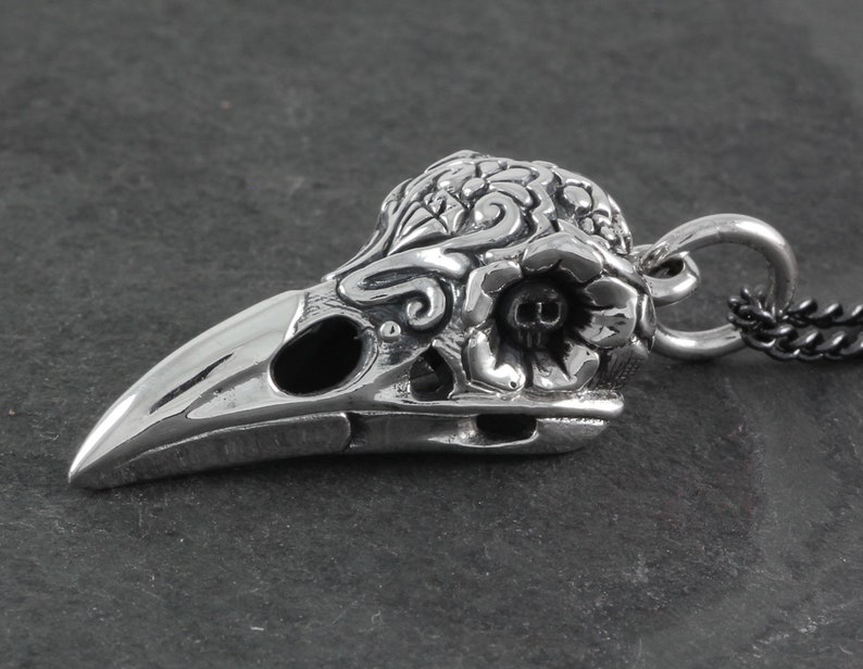 May include: Silver pendant featuring a detailed, ornate crow skull with floral designs. The skull has a small skull detail inside the eye socket. The pendant is attached to a black chain.