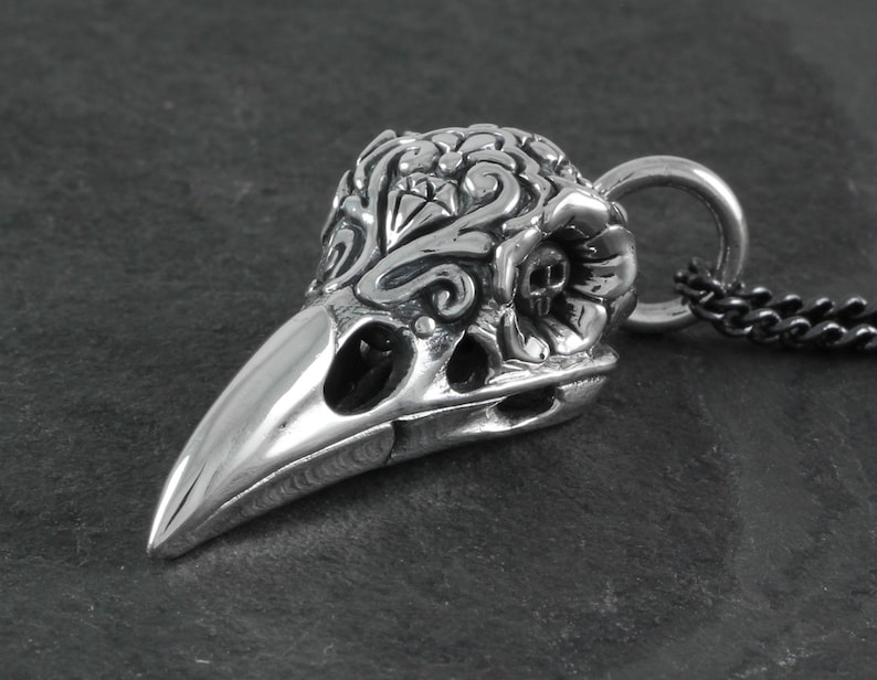 May include: A silver pendant in the shape of a crow skull with intricate floral designs. The pendant is attached to a black chain.