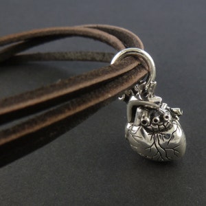 Pu&ograve; includere: Un braccialetto in corda di cuoio marrone con un ciondolo a forma di cuore anatomico argentato.