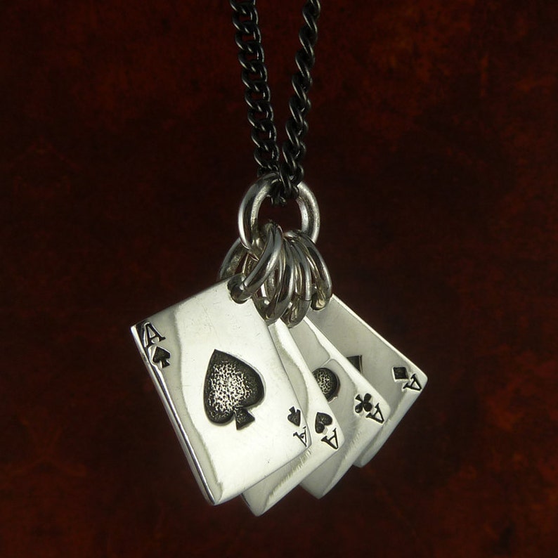Puede incluir: Un collar de plata con un colgante que presenta cuatro cartas de juego de as de picas. Las cartas est&aacute;n apiladas una encima de la otra y tienen una superficie texturizada. El collar est&aacute; en una cadena negra.