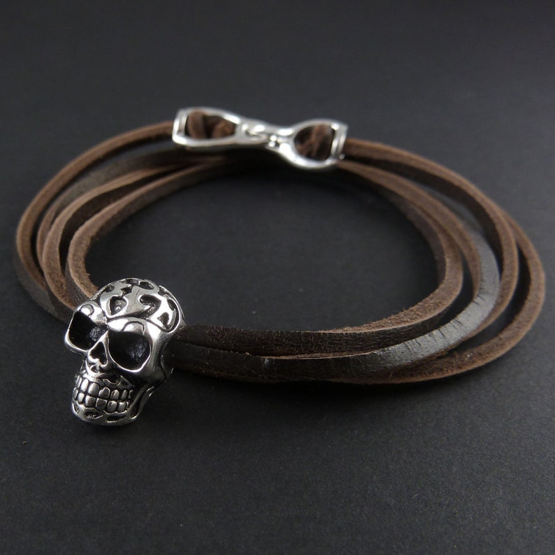 May include: A brown leather bracelet with a silver skull charm. The bracelet is made of three strands of leather and has a silver clasp.