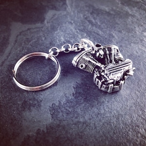 Puede incluir: Llavero de motor de motocicleta plateado. El llavero presenta un diseño de motor en miniatura detallado, conectado a una cadena y un llavero grande. El motor tiene detalles intrincados, incluidos cilindros y otros componentes mecánicos.