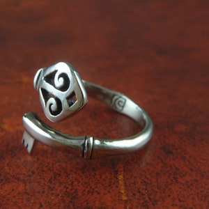 May include: A silver key ring with a detailed key design. The key is twisted and the ring is open. The ring is on a brown surface.