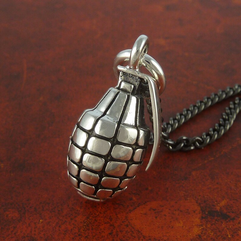 May include: A silver grenade pendant necklace with a black chain. The grenade is detailed with a textured surface and a pin.