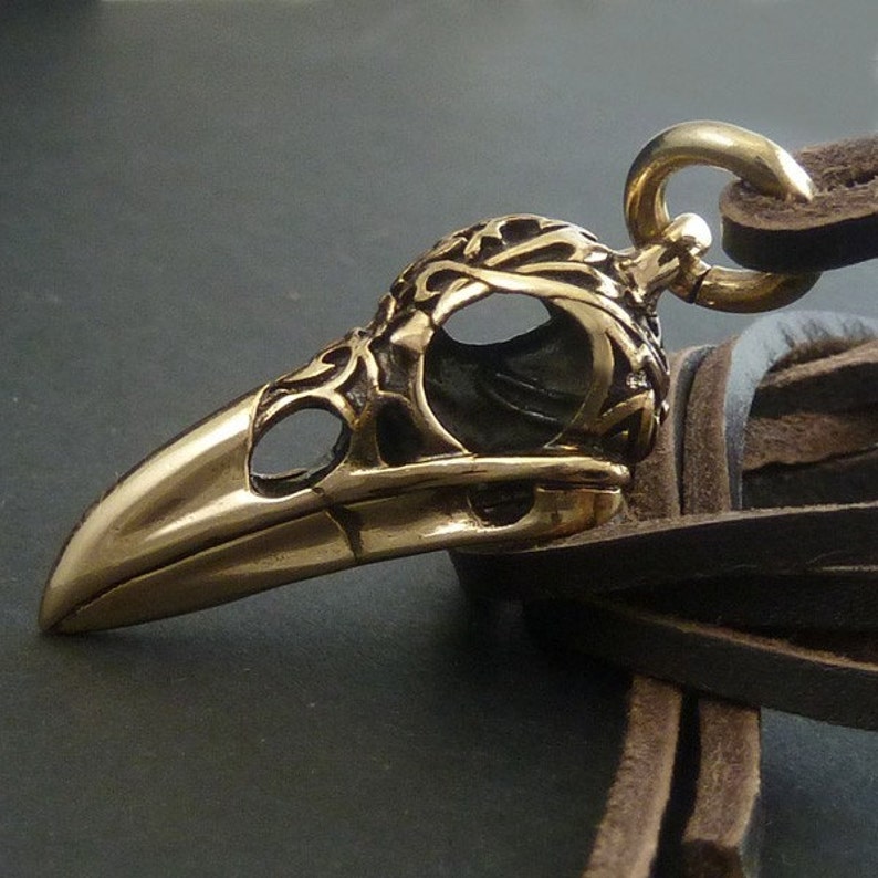 May include: A detailed bronze bird skull pendant with intricate carvings. The pendant is attached to a brown leather cord with a fringe.