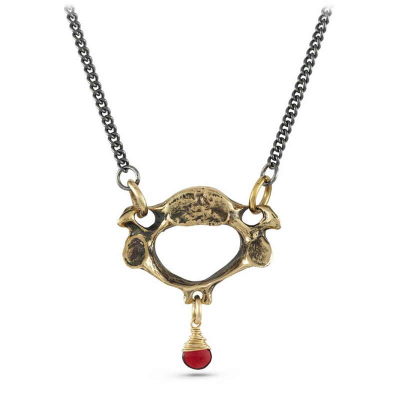 May include: A gold necklace with a pendant shaped like a human vertebra. The pendant has a small red gemstone hanging from it.