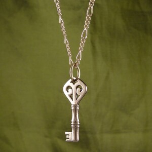 May include: A gold chain necklace with a silver key pendant. The key has an ornate design with a heart-shaped top.