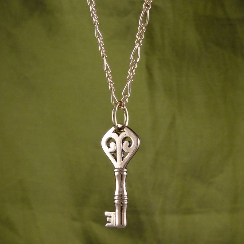 May include: A gold chain necklace with a silver key pendant. The key has an ornate design with a heart-shaped top.