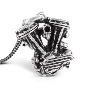 Puede incluir: Un collar con un colgante de plata que representa un motor de motocicleta en miniatura detallado. El colgante está unido a una cadena de plata.