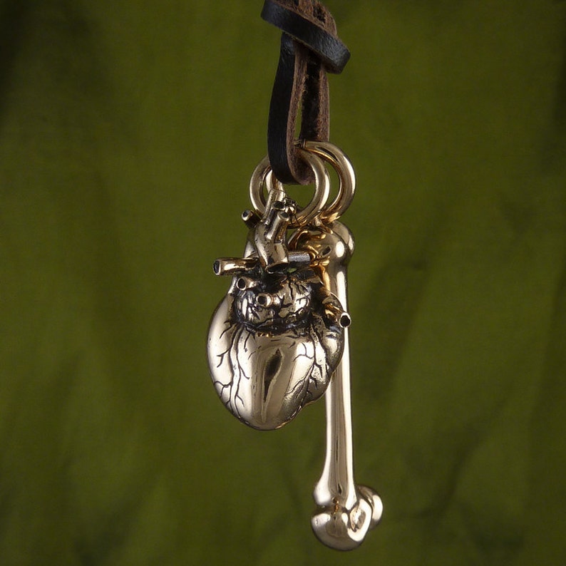May include: A gold-toned necklace with a detailed anatomical heart and bone pendant. The necklace is made of leather cord and has a simple design.