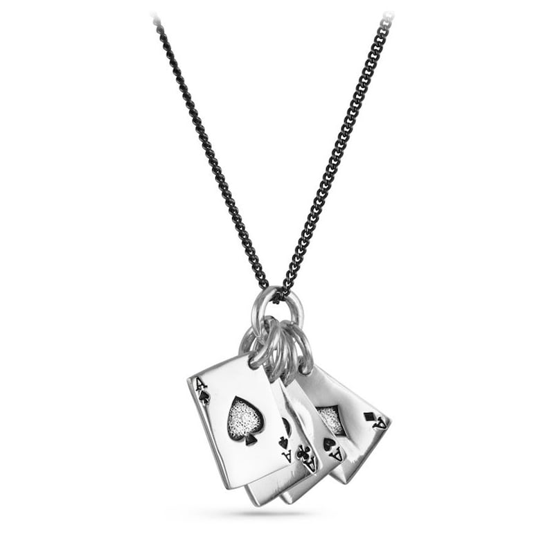 Puede incluir: Un collar de plata con una cadena negra y un colgante con cuatro dijes de cartas de juego de plata. Los dijes tienen forma de cartas de juego y muestran el as de picas, el as de diamantes, el as de tr&eacute;boles y el as de corazones.