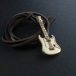 May include: A gold-colored electric guitar pendant on a brown leather cord. The guitar has a white body and a detailed design.