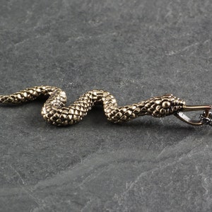 Puede incluir: Un collar con un colgante de serpiente de tono dorado con un diseño de serpiente texturizado y detallado. La serpiente está enroscada y tiene una pequeña boca abierta. El collar está unido a una cadena negra.