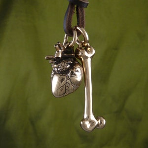 May include: A gold-toned necklace with a heart and bone pendant. The heart pendant is detailed with veins and the bone pendant is smooth. The necklace is on a brown leather cord.