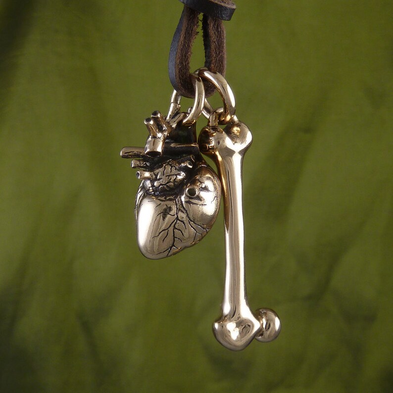 May include: A gold-toned necklace with a heart and bone pendant. The heart pendant is detailed with veins and the bone pendant is smooth. The necklace is on a brown leather cord.