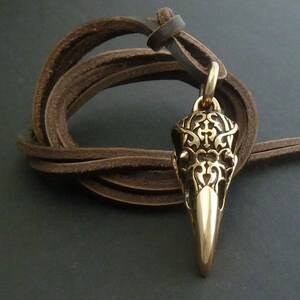 May include: A gold-toned pendant in the shape of a crow's skull with intricate details. The pendant is hanging from a brown leather cord.