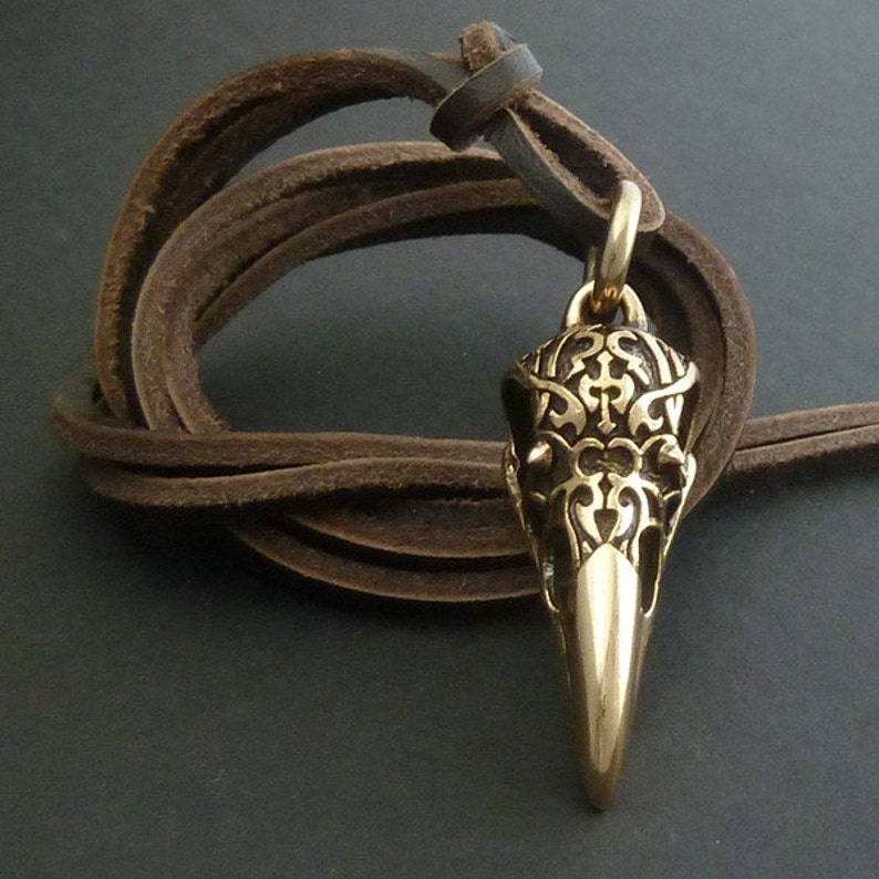 May include: A gold-toned pendant in the shape of a crow's skull with intricate details. The pendant is hanging from a brown leather cord.