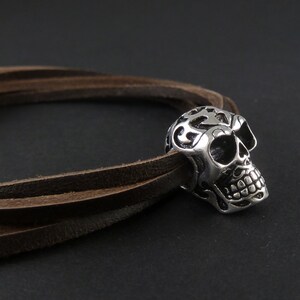 May include: A brown leather bracelet with a silver skull charm. The skull has intricate details and a floral design.
