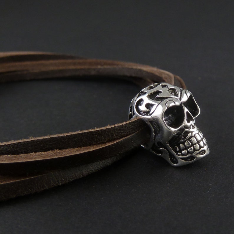 May include: A brown leather bracelet with a silver skull charm. The skull has intricate details and a floral design.