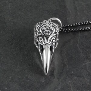 May include: A silver pendant necklace featuring a detailed, ornate skull design with a small diamond-shaped accent. The pendant is attached to a black chain.