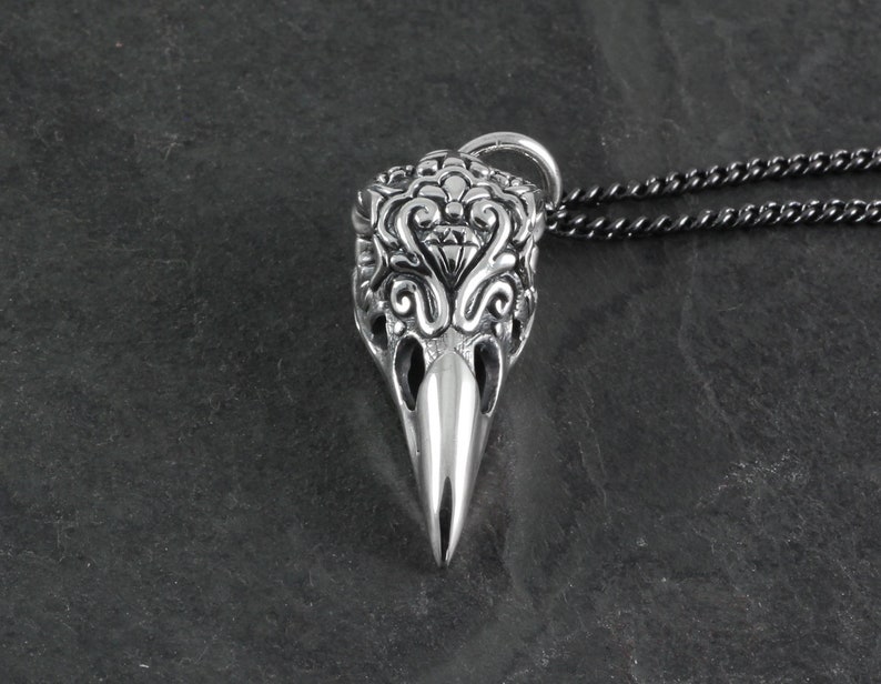 May include: A silver pendant necklace featuring a detailed, ornate skull design with a small diamond-shaped accent. The pendant is attached to a black chain.