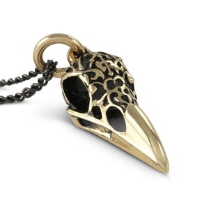 May include: Gold bird skull pendant necklace with black filigree detail. The pendant is attached to a black chain.