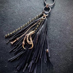May include: Black leather tassel keyring with gold-coloured chains and a bronze-toned bird claw charm. The keyring has a large silver-coloured ring and a smaller gold-coloured ring.