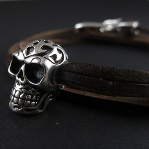 May include: A silver skull charm with intricate details on a brown leather bracelet.