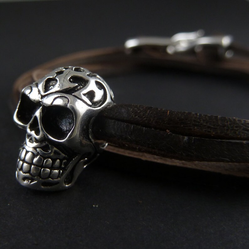 May include: A silver skull charm with intricate details on a brown leather bracelet.