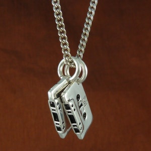 May include: A silver-colored necklace featuring a chain and two cassette tape pendants. The pendants have black details and are connected to the chain by a small ring. The background is a solid brown color.