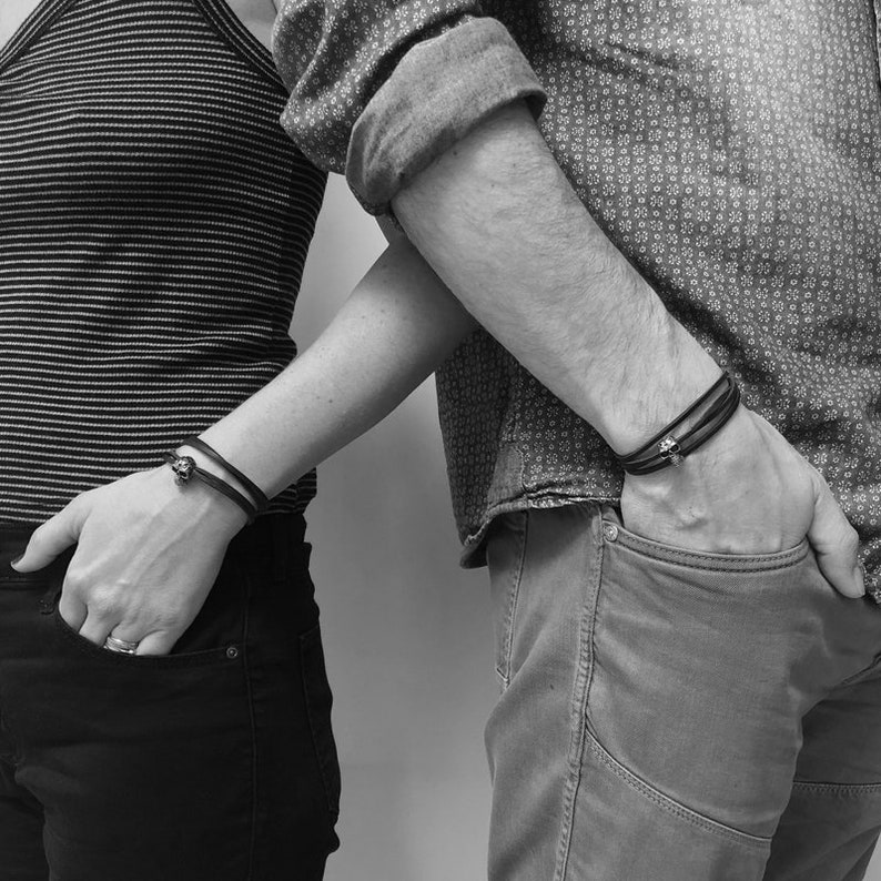 May include: Two people wearing black leather bracelets with silver skull charms. The bracelets are worn on the wrists of the couple.